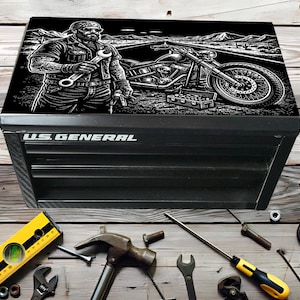 Peut inclure: Coffre à outils noir U.S. General avec un graphique noir et blanc d'un mécanicien et d'une moto sur le couvercle. Divers outils, dont un niveau, un marteau et des clés, sont éparpillés sur la surface en bois.