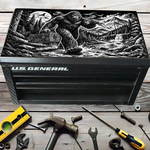 Peut inclure: Coffre à outils noir US General avec un motif Bigfoot sur le couvercle. Le motif est une illustration en noir et blanc de Bigfoot portant une clé et une boîte à outils dans un cadre forestier. Divers outils, dont un niveau, un marteau et des clés, sont éparpillés.