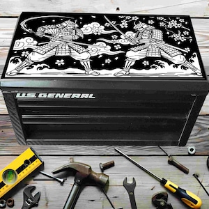 Puede incluir: Caja de herramientas negra con una tapa decorativa que muestra una ilustración en blanco y negro de dos guerreros samuráis. La caja tiene varios cajones y el texto "U.S. GENERAL" es visible. Varias herramientas están esparcidas alrededor de la caja.