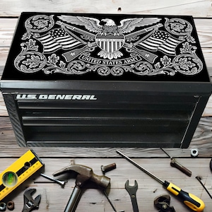 Puede incluir: Caja de herramientas negra US General con una tapa decorativa con un águila blanca, banderas y las palabras "United States Army". La caja está rodeada de varias herramientas, incluyendo un nivel, un martillo y llaves, sobre una superficie de madera.