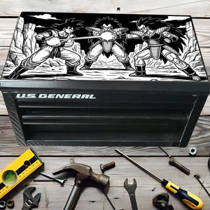 Könnte beinhalten: Schwarze US General Werkzeugkiste mit einer Schwarz-Weiß-Grafik von drei Anime-Charakteren. Die Werkzeugkiste ist von verschiedenen Werkzeugen umgeben, darunter eine Wasserwaage, ein Hammer und Schraubenschlüssel. Der Text "U.S. GENERAL" ist auf der Vorderseite der Werkzeugkiste aufgedruckt.