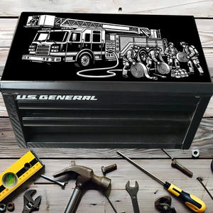 May include: Black U.S. General toolbox with a graphic of a fire truck and firefighters on the lid. The toolbox is surrounded by various tools, including a level, hammer, and wrenches. The toolbox has the words "U.S. GENERAL" in white.
