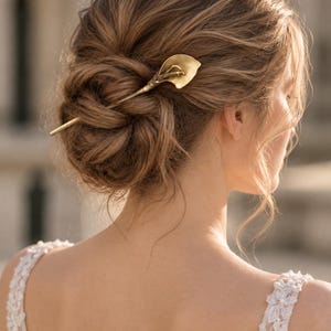 May include: A gold-toned metal hair pin shaped like a calla lily flower. The pin features a long, slender stem that secures into a styled updo, with the curved, trumpet-shaped petal of the lily resting against the hair to serve as a decorative accent.