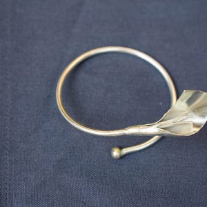 Peut inclure: Un rond de serviette argenté avec un motif floral. L'anneau est circulaire avec un élément stylisé en forme de feuille. Le métal a une finition polie et la pièce repose sur un tissu bleu foncé.