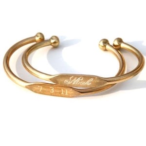 May include: Two gold-colored cuff bracelets. One bracelet is engraved with the name "Noah", and the other is engraved with the date "7-3-11". Each bracelet has a rounded ball at each end.