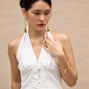 May include: A set of gold-toned jewelry featuring calla lily motifs, including a pair of long drop earrings, a hair accessory, a ring, and an open cuff bracelet. The jewelry is worn with a white sleeveless halter top with a button-down front.
