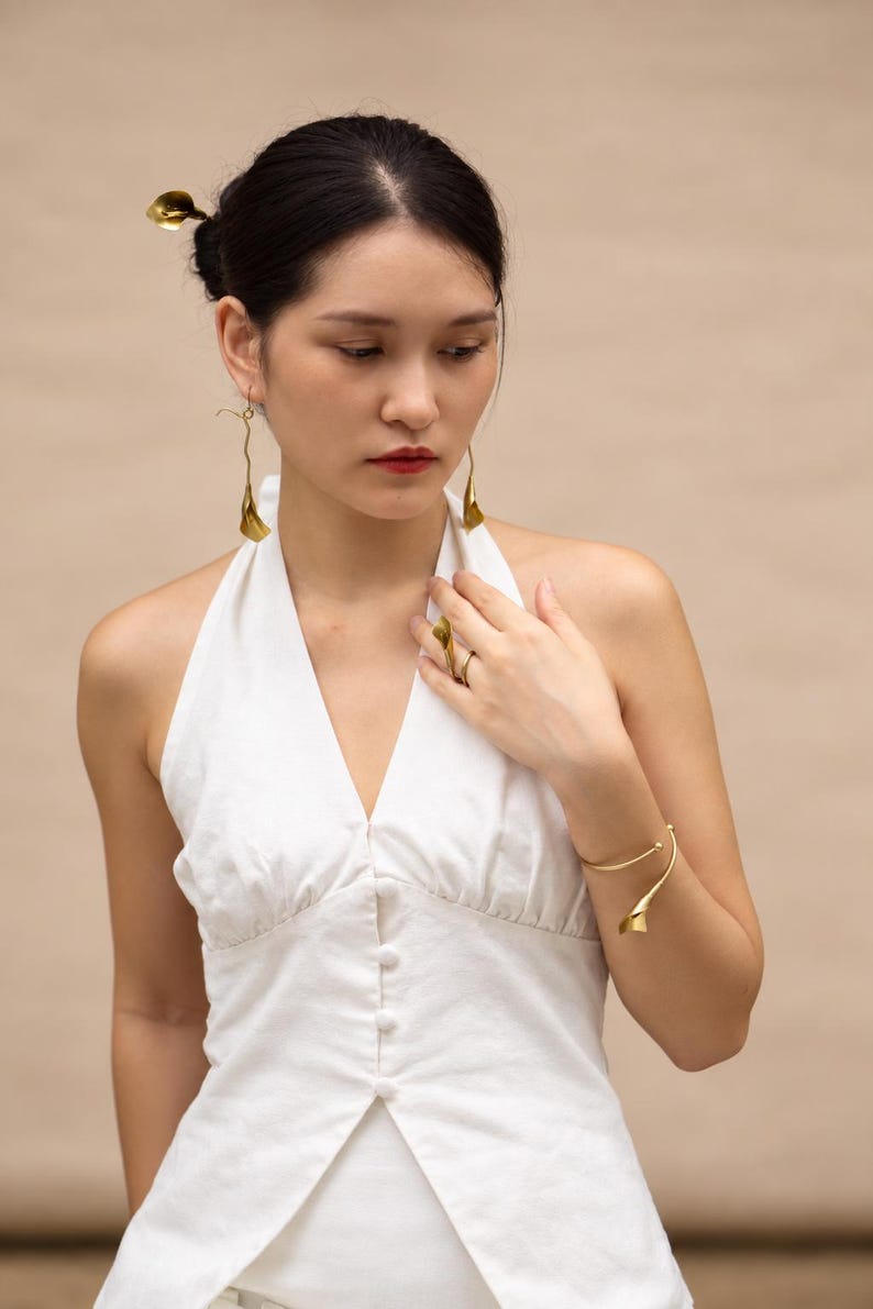 May include: A set of gold-toned jewelry featuring calla lily motifs, including a pair of long drop earrings, a hair accessory, a ring, and an open cuff bracelet. The jewelry is worn with a white sleeveless halter top with a button-down front.