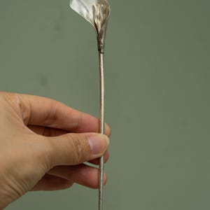 May include: A silver-colored metal hair stick featuring a sculpted calla lily flower at the top. The long, slender, straight stem extends downward from the base of the flower, which has a smooth, curved petal shape and a central stamen detail.