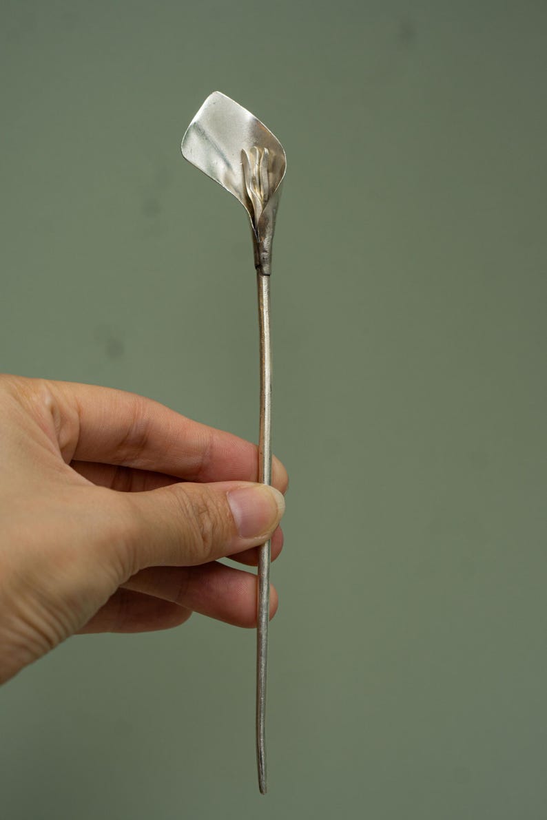 May include: A silver-colored metal hair stick featuring a sculpted calla lily flower at the top. The long, slender, straight stem extends downward from the base of the flower, which has a smooth, curved petal shape and a central stamen detail.