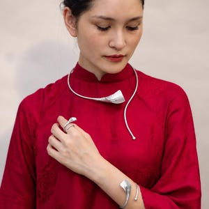 May include: A silver jewelry set consisting of a collar necklace, a cuff bracelet, a ring, and a hair accessory, each featuring a stylized calla lily motif. The pieces are worn with a solid red long-sleeved garment with a high collar and subtle texture.