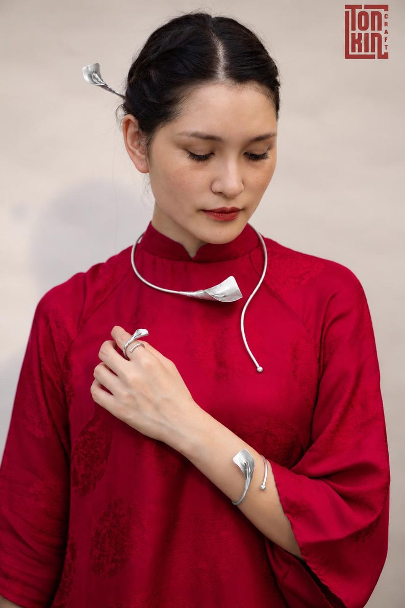 May include: A silver jewelry set consisting of a collar necklace, a cuff bracelet, a ring, and a hair accessory, each featuring a stylized calla lily motif. The pieces are worn with a solid red long-sleeved garment with a high collar and subtle texture.