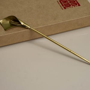 May include: A gold-toned metal hair pin featuring a stylized calla lily flower at one end. The long, slender, pointed stem extends from the base of the flower. The hair accessory is crafted with a smooth, polished finish and a metallic sheen.