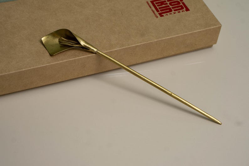 May include: A gold-toned metal hair pin featuring a stylized calla lily flower at one end. The long, slender, pointed stem extends from the base of the flower. The hair accessory is crafted with a smooth, polished finish and a metallic sheen.