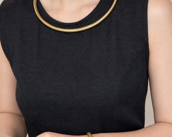 May include: A smooth, matte gold-toned metal choker necklace and a matching circular bangle bracelet. Both pieces feature a simple, rounded, solid design with a consistent metallic finish, worn against a plain black sleeveless garment.