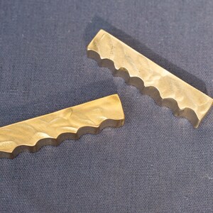 May include: Two brass-colored, rectangular drawer pulls with a wavy edge. The pulls are lying on a dark blue fabric surface. The pulls have a textured surface and are likely for a cabinet or drawer.