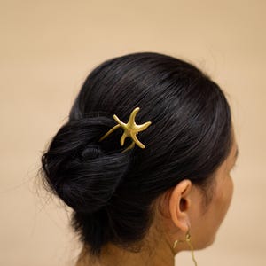 Op de afbeelding: Een goudkleurige zeesterren haarspeld is in een knot kapsel geplaatst. Het haar is zwart en de achtergrond is effen beige. Het haaraccessoire is een decoratief item.