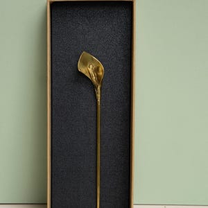 May include: A gold-colored metal hair pin shaped like a calla lily flower, featuring a long, slender stem and a curved petal head. The hair pin is presented inside a rectangular cardboard box with a black textured foam insert.