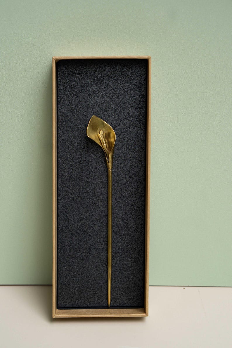 May include: A gold-colored metal hair pin shaped like a calla lily flower, featuring a long, slender stem and a curved petal head. The hair pin is presented inside a rectangular cardboard box with a black textured foam insert.