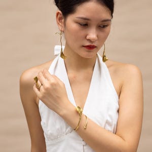 May include: A set of gold-toned jewelry featuring a calla lily motif, including a pair of long drop earrings, a hair pin, a cuff bracelet with a floral end, and a matching ring, all displayed on a white sleeveless button-down top.
