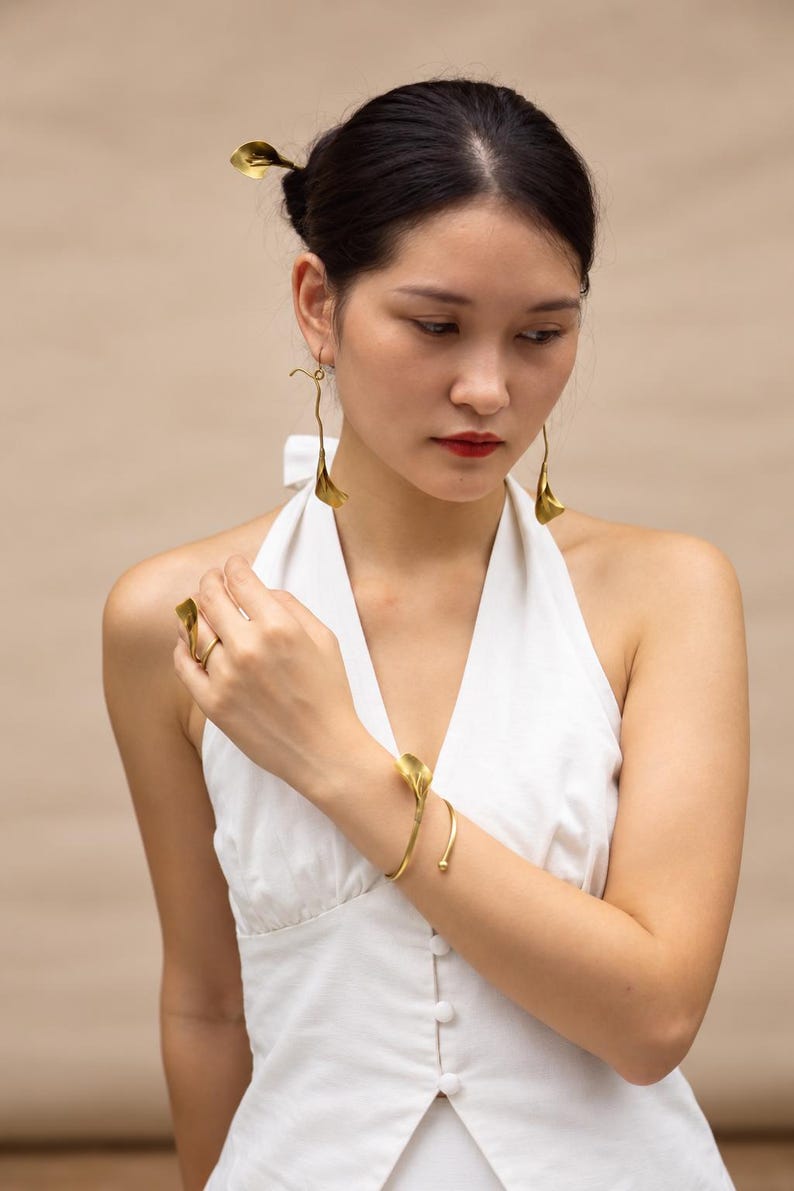 May include: A set of gold-toned jewelry featuring a calla lily motif, including a pair of long drop earrings, a hair pin, a cuff bracelet with a floral end, and a matching ring, all displayed on a white sleeveless button-down top.