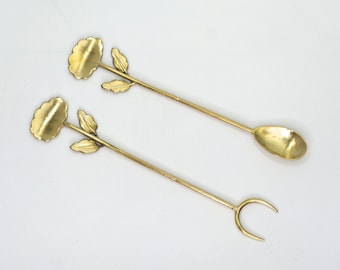 May include: Two gold-toned metal spoons with floral designs. The spoons have a flower-shaped handle and a leaf design on the stem. One spoon has a rounded spoon head, and the other has a fork-like head.