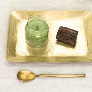 Könnte beinhalten: Ein goldfarbenes Tablett mit gehämmerter Oberfläche, auf dem eine grüne Glaskerze und ein Stück Treibholz liegen. Ein goldfarbener Löffel ist ebenfalls enthalten.