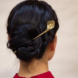 May include: A gold-toned metal hair pin shaped like a calla lily flower, inserted into a dark-haired bun. The hair accessory features a slender stem and a curved, petal-like top, providing a metallic accent to the dark hair styled in a low bun.