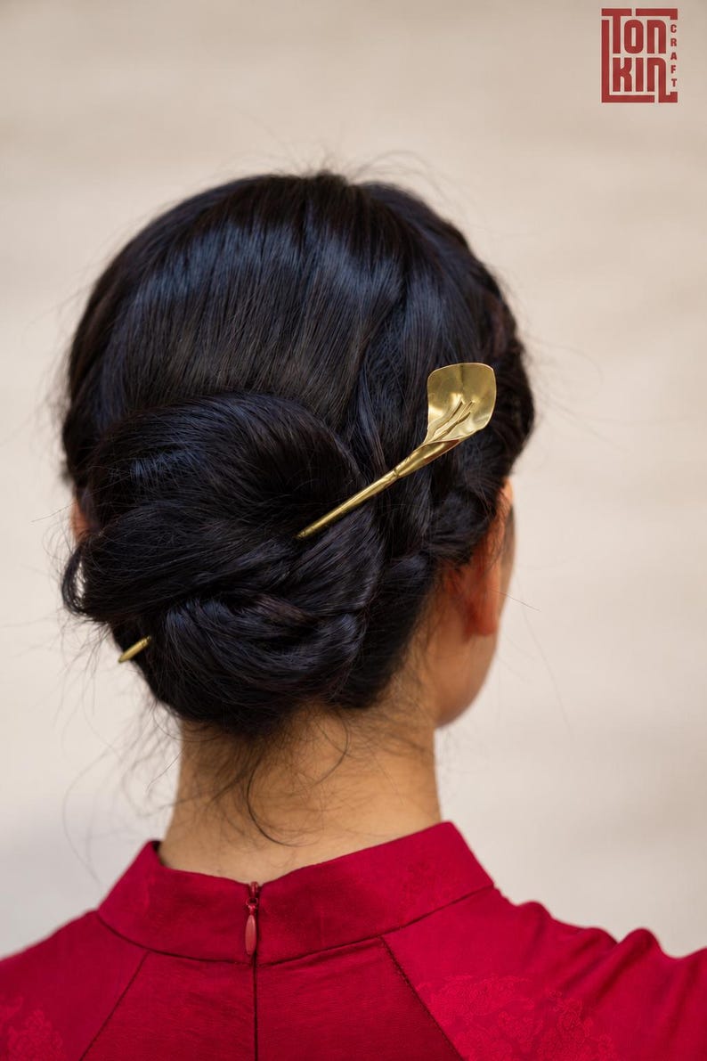 May include: A gold-toned metal hair pin shaped like a calla lily flower, inserted into a dark-haired bun. The hair accessory features a slender stem and a curved, petal-like top, providing a metallic accent to the dark hair styled in a low bun.