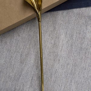 May include: A gold-toned metal hair stick featuring a sculpted calla lily flower at the top. The accessory has a long, slender, straight stem that tapers to a point at the bottom, with a smooth, polished finish throughout the entire piece.