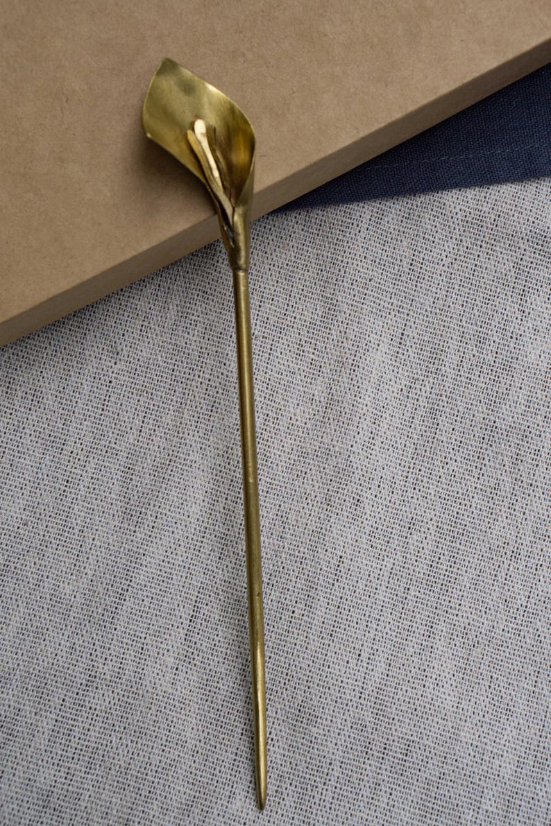 May include: A gold-toned metal hair stick featuring a sculpted calla lily flower at the top. The accessory has a long, slender, straight stem that tapers to a point at the bottom, with a smooth, polished finish throughout the entire piece.