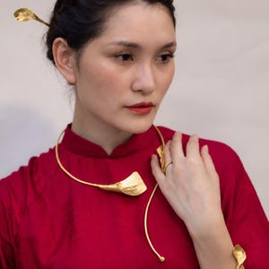 May include: A gold-toned metal jewelry set consisting of a collar necklace, a bracelet, a hair pin, and a ring, each featuring a stylized leaf or petal motif. The pieces are worn with a solid red high-neck garment. The Tonkin Craft logo is visible.