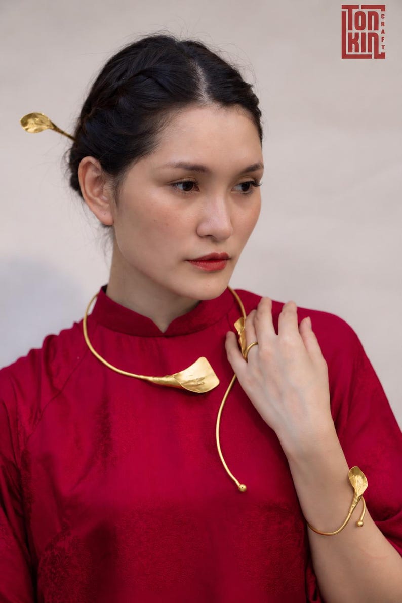 May include: A gold-toned metal jewelry set consisting of a collar necklace, a bracelet, a hair pin, and a ring, each featuring a stylized leaf or petal motif. The pieces are worn with a solid red high-neck garment. The Tonkin Craft logo is visible.