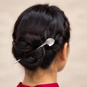 May include: A silver-toned hair pin featuring a calla lily design inserted into a dark brown braided bun hairstyle. The metal pin has a smooth, matte finish and a slender, tapered shaft that secures the hair in place with a floral-shaped topper.