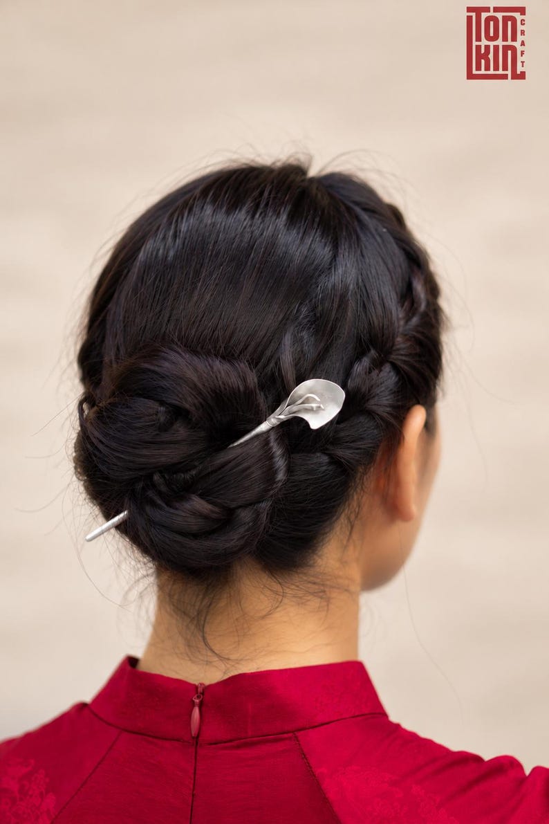 May include: A silver-toned hair pin featuring a calla lily design inserted into a dark brown braided bun hairstyle. The metal pin has a smooth, matte finish and a slender, tapered shaft that secures the hair in place with a floral-shaped topper.