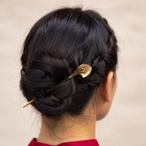 May include: A gold-toned metal hair pin featuring a stylized calla lily design at one end. The hair pin is inserted into a dark brown braided bun hairstyle. The accessory has a smooth, metallic finish and a slender, tapered shaft design.