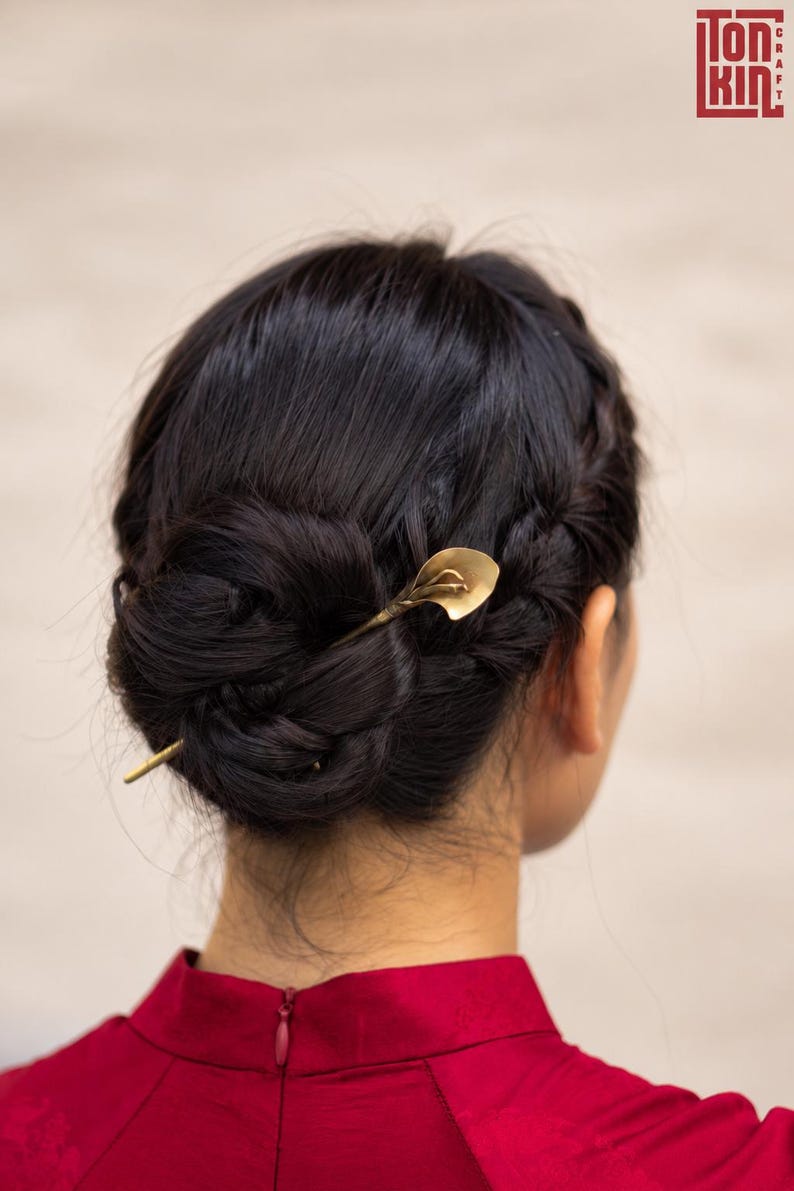 May include: A gold-toned metal hair pin featuring a stylized calla lily design at one end. The hair pin is inserted into a dark brown braided bun hairstyle. The accessory has a smooth, metallic finish and a slender, tapered shaft design.