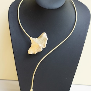 May include: A gold-colored necklace featuring a ginkgo leaf pendant. The pendant has detailed veining and is attached to a slender, curved chain. The necklace is displayed on a black jewelry stand.