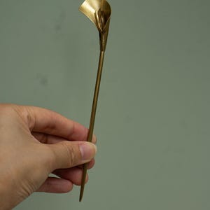 May include: A gold-toned metal hair stick featuring a sculpted calla lily flower at the top. The accessory has a long, slender, straight stem that tapers to a point at the bottom, with a smooth, polished finish throughout the entire piece.