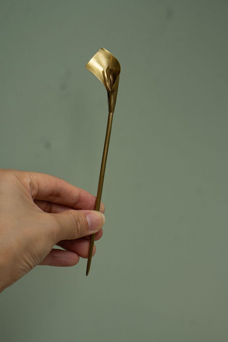 May include: A gold-toned metal hair stick featuring a sculpted calla lily flower at the top. The accessory has a long, slender, straight stem that tapers to a point at the bottom, with a smooth, polished finish throughout the entire piece.