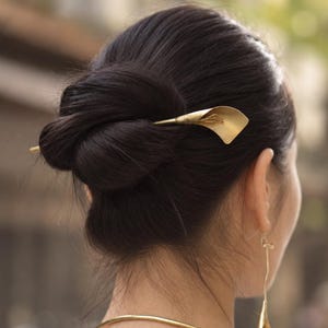May include: A gold-toned hair pin shaped like a calla lily flower, secured in a dark hair bun. The set includes a matching gold-toned drop earring featuring a small calla lily pendant and a simple, thin gold-toned metal neck collar.
