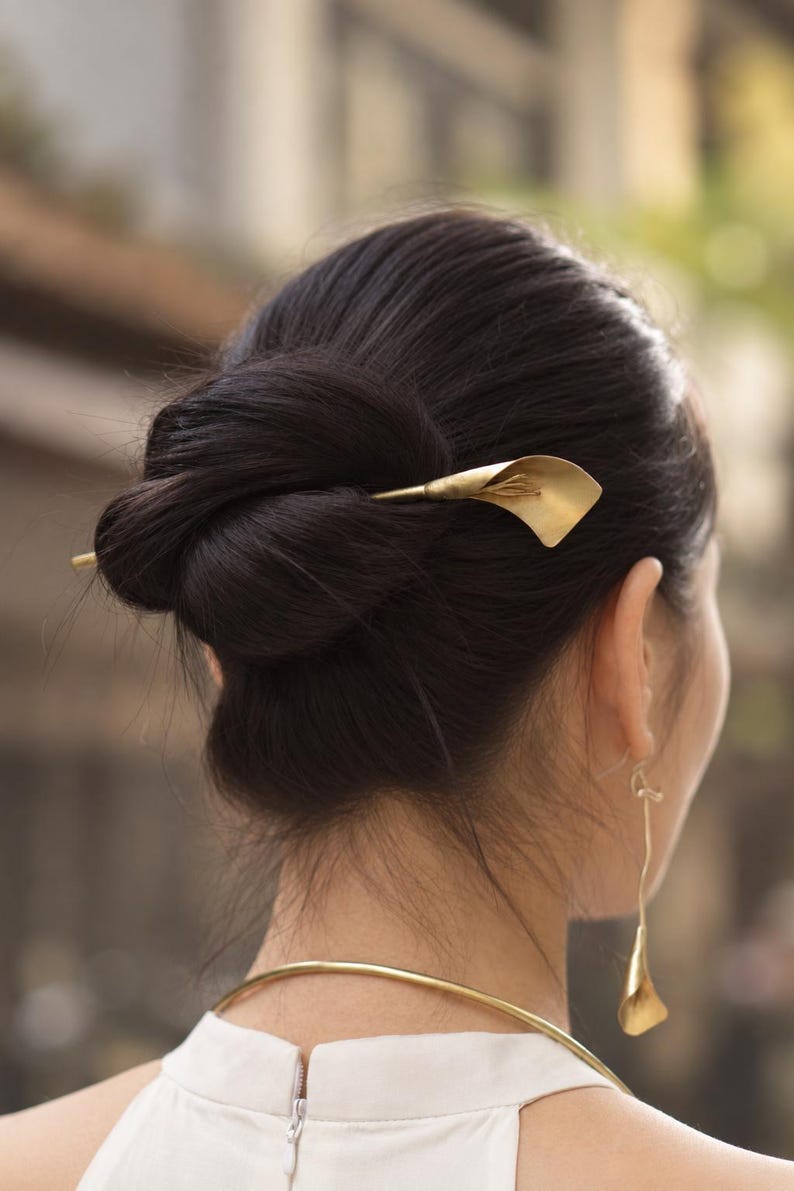 May include: A gold-toned hair pin shaped like a calla lily flower, secured in a dark hair bun. The set includes a matching gold-toned drop earring featuring a small calla lily pendant and a simple, thin gold-toned metal neck collar.
