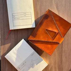 Pu&ograve; includere: Due taccuini MD Paper color bianco sporco e una copertina in pelle marrone chiaro. Il taccuino in alto presenta un'etichetta con il testo "VOLUME," "START," "FINISH," e "FROM THE LIBRARY OF." Il taccuino in basso mostra un disegno di una mano.
