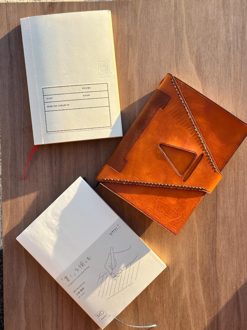 Pu&ograve; includere: Due taccuini MD Paper color bianco sporco e una copertina in pelle marrone chiaro. Il taccuino in alto presenta un'etichetta con il testo "VOLUME," "START," "FINISH," e "FROM THE LIBRARY OF." Il taccuino in basso mostra un disegno di una mano.