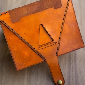 Pu&ograve; includere: Una custodia in pelle color cuoio a forma di busta con una patta triangolare dotata di un intaglio centrale a forma di cono. I bordi sono rifiniti con cuciture scure e la patta &egrave; chiusa da un bottone a pressione in metallo color ottone.