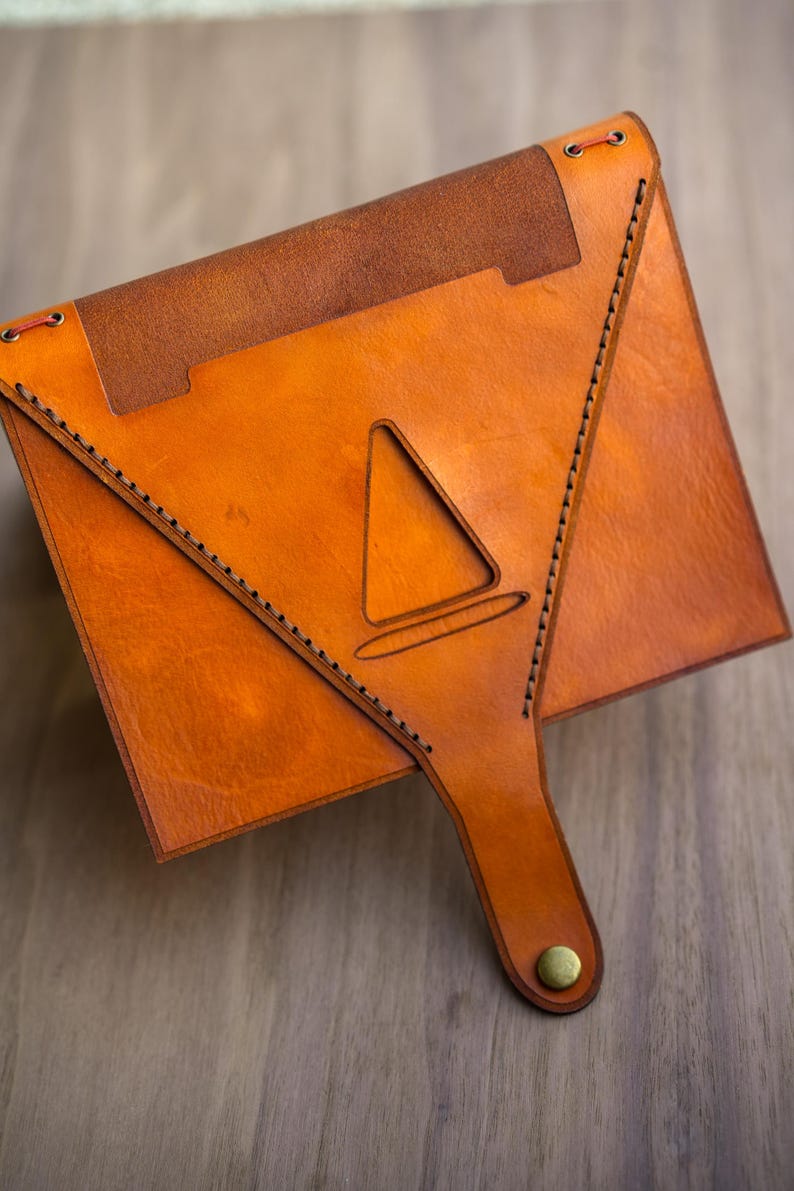 Pu&ograve; includere: Una custodia in pelle color cuoio a forma di busta con una patta triangolare dotata di un intaglio centrale a forma di cono. I bordi sono rifiniti con cuciture scure e la patta &egrave; chiusa da un bottone a pressione in metallo color ottone.