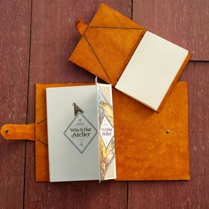 Pu&ograve; includere: Due copertine in pelle color cuoio per libri o diari, una aperta e una chiusa. La copertina aperta contiene una copia di Witch Hat Atelier volume 4, con un logo a forma di diamante e un'illustrazione grafica sulla pagina interna.