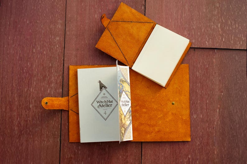 Pu&ograve; includere: Due copertine in pelle color cuoio per libri o diari, una aperta e una chiusa. La copertina aperta contiene una copia di Witch Hat Atelier volume 4, con un logo a forma di diamante e un'illustrazione grafica sulla pagina interna.