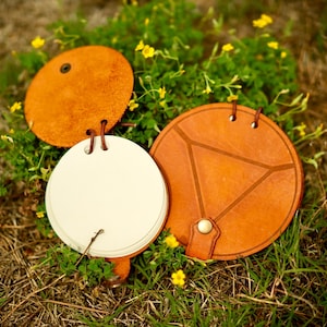 May include: A set of three leather-bound notebooks in a natural tan color. One notebook is open, revealing white pages. The other two are closed, with one featuring a geometric design. The notebooks are resting on grass with yellow flowers.
