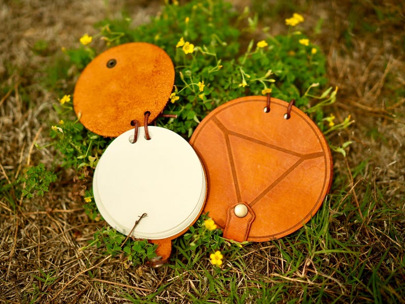 May include: A set of three leather-bound notebooks in a natural tan color. One notebook is open, revealing white pages. The other two are closed, with one featuring a geometric design. The notebooks are resting on grass with yellow flowers.