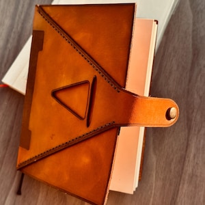 Pu&ograve; includere: Un diario in pelle color cuoio con un design a intaglio triangolare sulla copertina anteriore. La copertina &egrave; fissata con un cinturino in pelle abbinato che include una chiusura a bottone automatico in metallo. Il diario contiene pagine bianche.